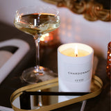 Brennende CUVÉE Duftkerze Chardonnay auf einem goldenen Tablett neben einem Glas Champagner