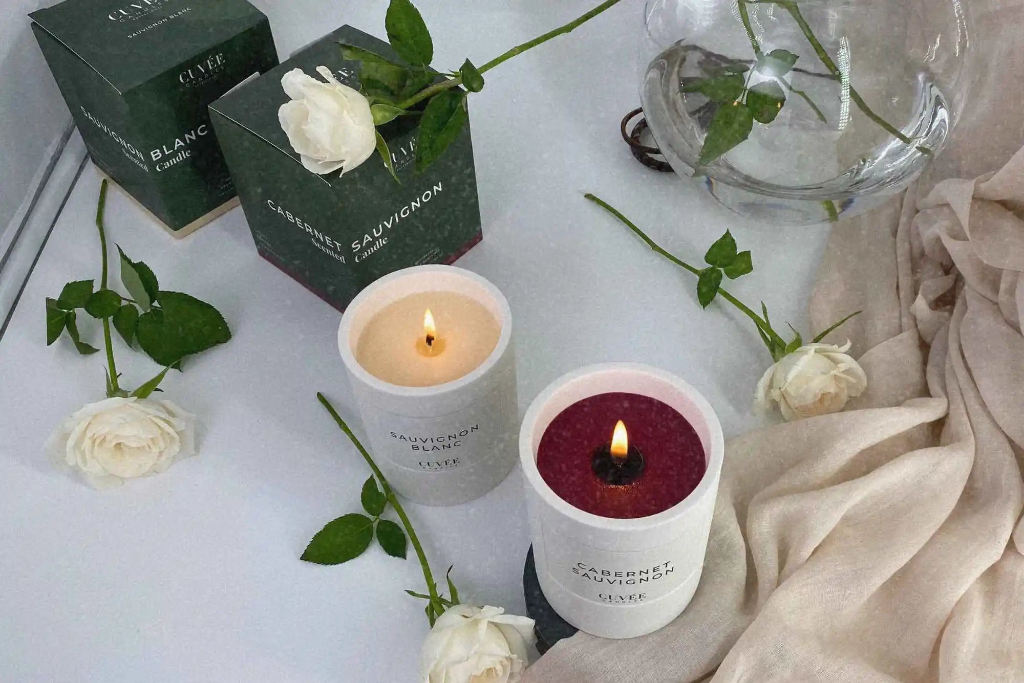 Edle weiße Rosen und edle, grüne CUVÉE CANDLES Kerzenschachtel neben luxuriöser, weißer Sauvignon Blanc Duftkerze und brennender Cabernet Sauvingon Duftkerze von CUVÉE CANDLES als einzigartige Erinnerung an den Hochzeitstag.