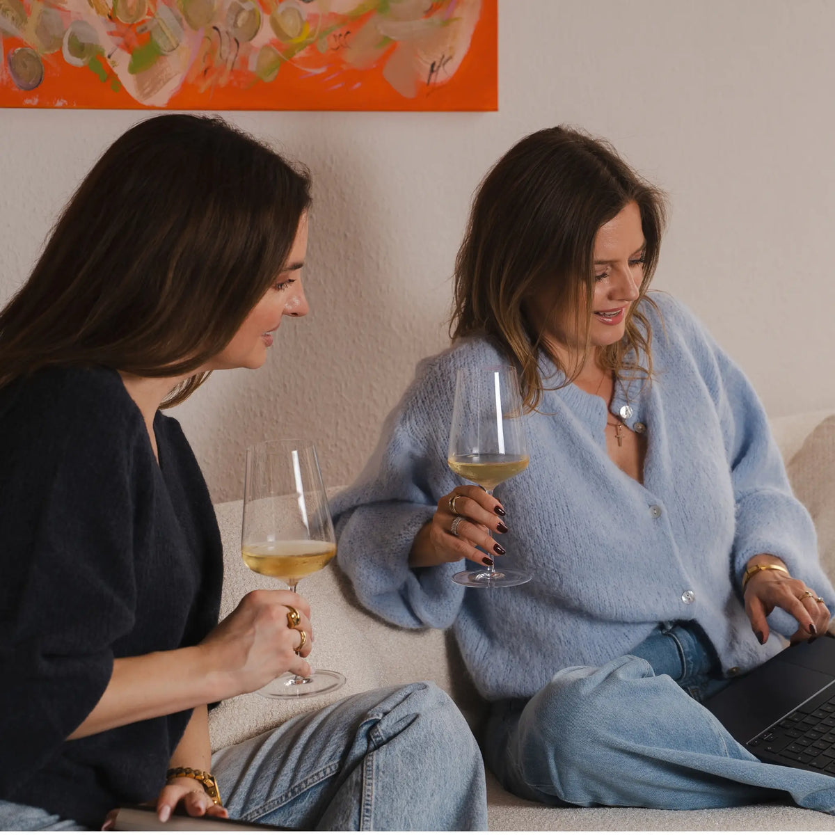 Die beiden Gründerinnen mit Weißwein auf dem Sofa, eine arbeitet am Laptop, dahinter ein orangefarbenes Gemälde
