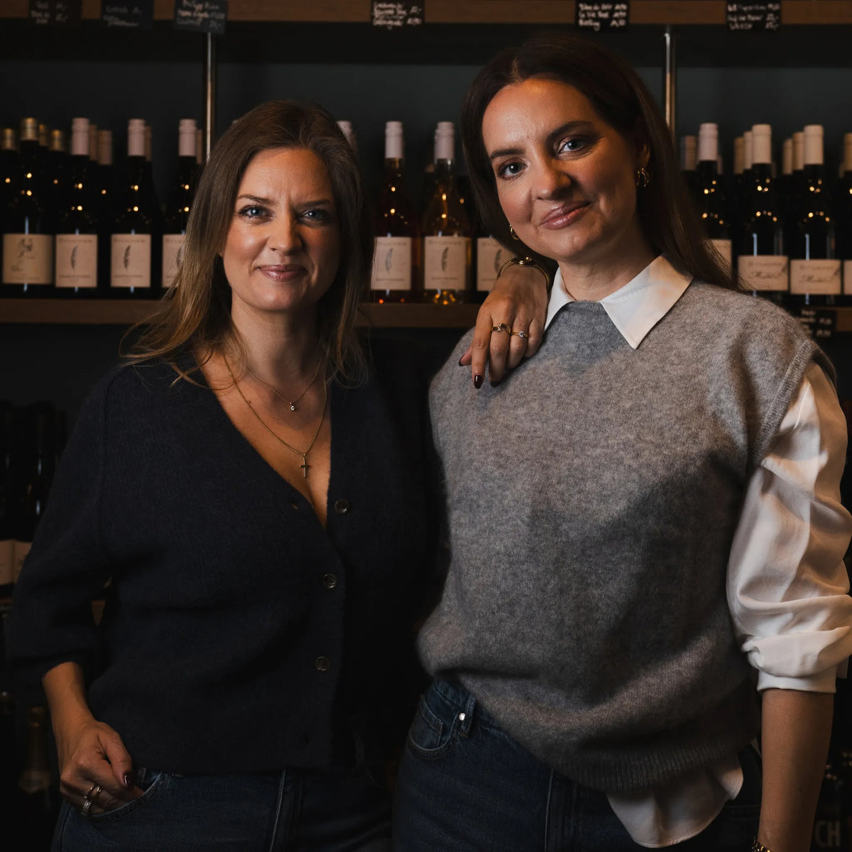 Die Gründerinnen von CUVÉE CANDLES lachen sich vor einem Weinregal an, eine lehnt den Arm auf die Schulter der anderen